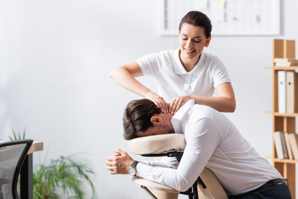 Happy masseuse doing neck massage for businessman in office on blurred background